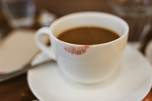 Close up of red lipstick smudges on hot latte in white coffee cup on wood table near window in coffee shop in the morning in vintage soft tone, metaphor of female office worker.