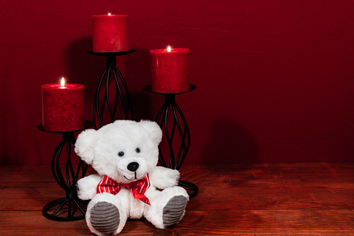 Three red candles in metal holders and red rose, one teddy bear on wooden table.