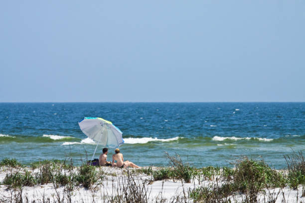 Couple relaxing on the beach at spring in ship island Taken this picture of a couple at a distant relaxing on the beach of ship island off gulf of mexico. The umbrella is tilted due to strong wind and the waves. mississippi delta stock pictures, royalty-free photos & images