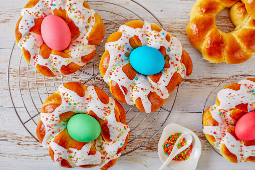 homemade sweet Italian Easter Bread Rings glazed around dyed egg and topped with colorful sprinkles on a wire rack, on an old white wooden table, view from above, flatlay