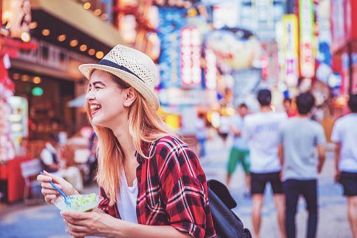Travel, Tourism, Vacation, Enjoyment - Young attractive cheerful female tourist holding a cup of ice cream while having a cheerful conversation with her friends at the famous market streets of Shinsekai, in Osaka, Japan