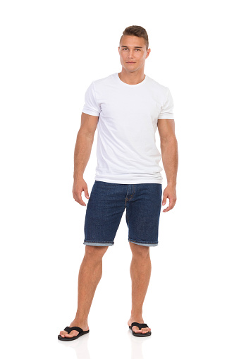 Handsome young man in white shirt, jeans shorts and black sandals is standing and looking at camera. Front view. Full length studio shot isolated on white.