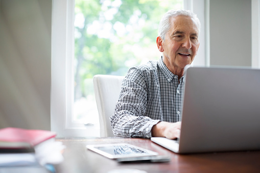 Smiling senior man surfing on laptop at desk against window. Retired male is sitting in room. He is in formalwear.
