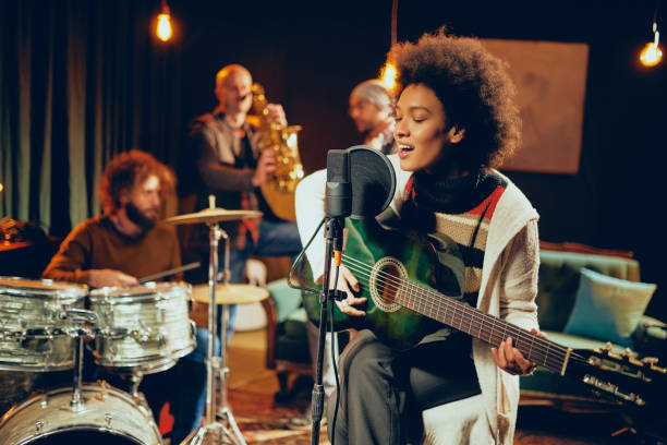 Mixed race woman singing and playing guitar. Mixed race woman singing and playing guitar while sitting on chair with legs crossed. In background drummer, saxophonist and bass guitarist. recording studio stock pictures, royalty-free photos & images