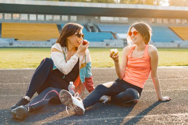 mode de vie sain et une alimentation saine concept. sourire, mère de remise en forme et teen fille ensemble eating apple assis sur le stade après la formation le jour de printemps ensoleillé - sport food exercising eating photos et images de collection