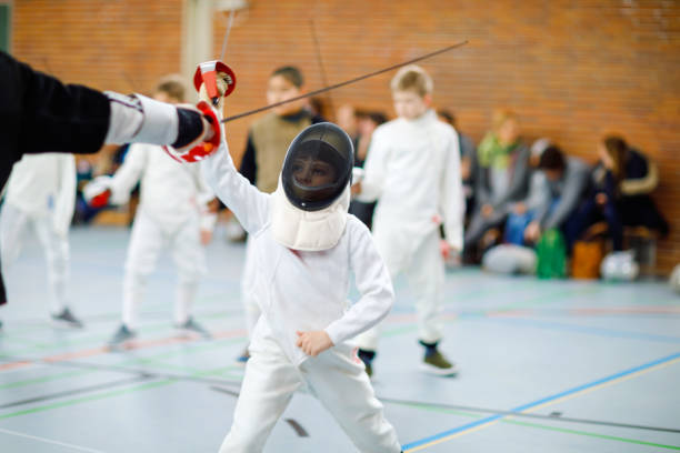 Little kid boy fencing on a fence competition. Child in white fencer uniform with mask and sabre. Active kid training with teacher and children. Healthy sports and leisure. stock photo