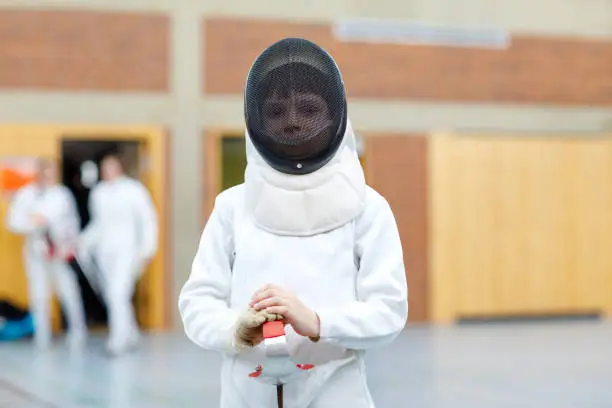 Little kid boy fencing on a fence competition. Child in white fencer uniform with mask and sabre. Active kid training with teacher and children. Healthy sports and leisure