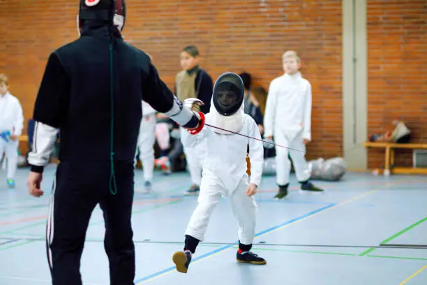 Little kid boy fencing on a fence competition. Child in white fencer uniform with mask and saber. Active kid training with teacher and children. Healthy sports and leisure