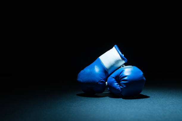 A pair of blue boxing gloves sit on a black background with a light shining from overhead.