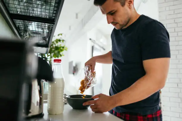 Young man preparing cornflakes and milk for breakfast in the kitchen of his apartment