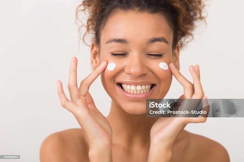 Laughing girl applying moisturizing cream on her face Skin care. Laughing girl applying moisturizing cream on her face over white background Moisturizer Stock Photo