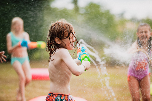 A shot of young children in a garden, they are having fun together enjoying a water gun fight with colorful water guns and a garden hose.