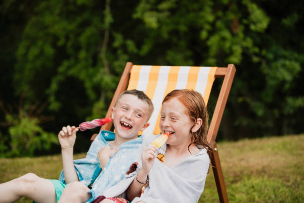 Boys Enjoying Summer A front view shot of two young boys sitting together enjoying popsicles, they are wearing towels and laughing and smiling. popsicle stick stock pictures, royalty-free photos & images