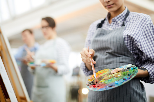 Mid section portrait of unrecognizable female artist holding palette mixing oil colors while painting picture on easel in art studio, copy space