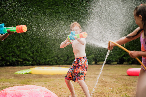 A front view shot of young children in a garden, they are having fun together enjoying a water fight with a garden hose and colorful water guns.