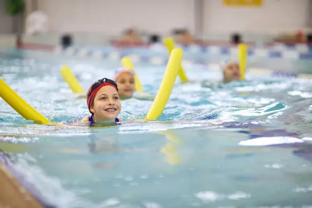 Happy little girl in swimming cap learning to swim with swim noodle at the lesson in pool