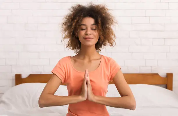 African-american woman sitting in namaste yoga position and relaxing on bed in the morning