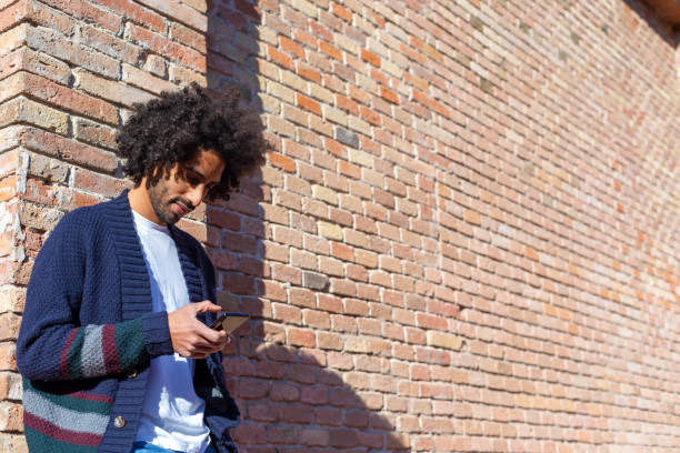 istock Young handsome African man using his smartphone with smile while leaning on a bricked wall outdoors in sunny day 1125927010