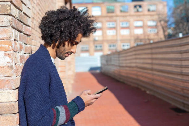 istock Young handsome African man using his smartphone with smile while leaning on a bricked wall outdoors in sunny day 1125927019
