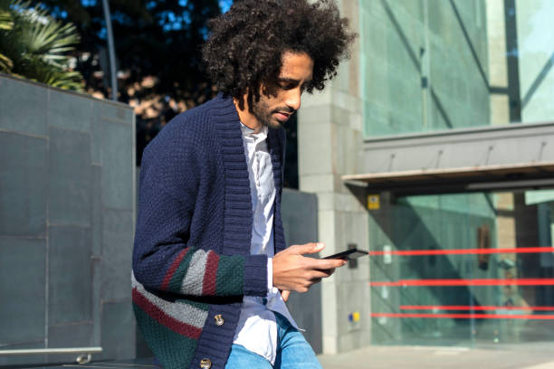 istock Young handsome African man using his smartphone with smile while sitting on a bench outdoors in sunny day 1125927343