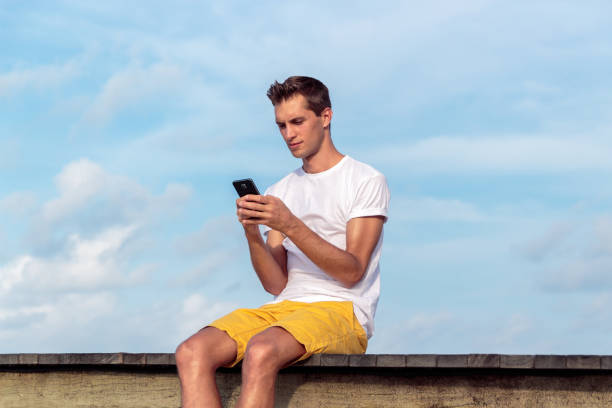 istock Man seated on a pier in a tropical location using his smartphone. Sky with clouds as background 1125937980