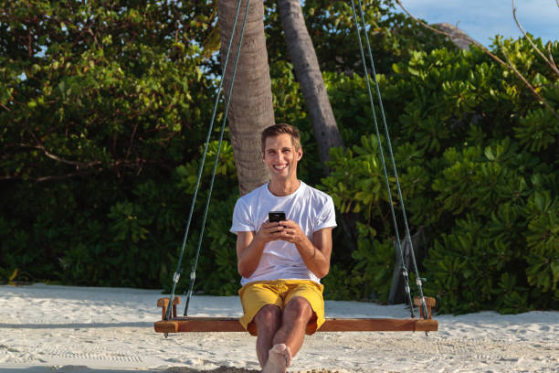 istock young happy man seated on a swing and using his phone. White sand and jungle as background 1125938094