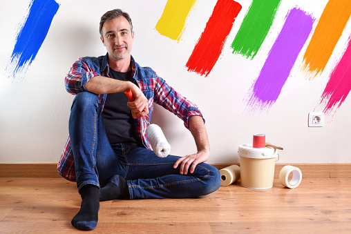 Man sitting on a parquet floor prepared with paint material to repaint his house and wall painted with seven colors. Front view. Horizontal composition.