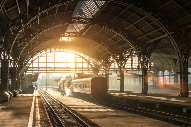 Old railway station with a train and a locomotive on the platform awaiting departure. Evening sunshine rays in smoke arches