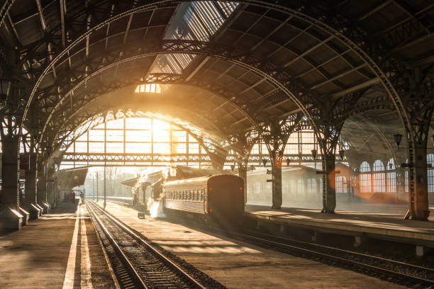 antiga estação ferroviária com um trem e uma locomotiva na plataforma aguardando a partida. raios de luz do sol em arcos fumos a noite. - estação de trem - fotografias e filmes do acervo