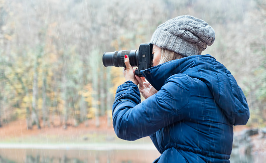 Woman taking photo with full frame camera in woods on rainy day - winter clothes and blue coat on