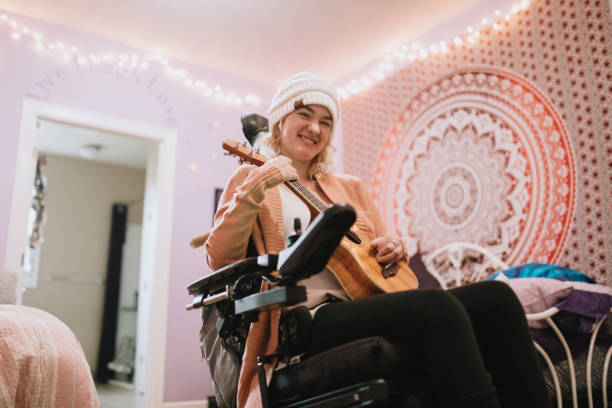 Cheerful Young Woman In Wheelchair At Home With Ukulele A independent  young adult woman with cerebral palsy going about some of her daily routines at home.  She shows off her ukulele guitar playing skills. physical disability photos stock pictures, royalty-free photos & images