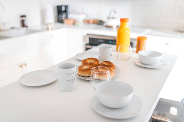 Close-up of tableware and breads on kitchen island. Mugs with bowls and plates are arranged on counter. Interior of brightly lit kitchen.