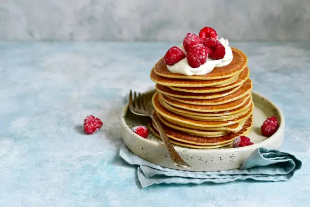 Stack of homemade delicious hot pancakes with honey, cream and raspberry on a plate on a light slate, stone or concrete background.