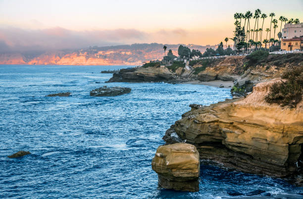 La Jolla Cove, San Diego, CA, USA - September 27, 2023.  The young woman lounges casually on a jagged rock, her posture relaxed and a mobile phone in her hand. Her hair is pulled back in a messy ponytail and she wears a green sweater and shorts. She seems both calm and indifferent to the stormy sea below. Waves crash against the rocky shore, sending sprays of saltwater into the air. She appears so tiny compared to the vast expanse of the ocean that stretches out before her.