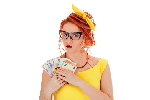 Greedy woman holding money, euro cash refusing to give it. Caucasian person in yellow dress and retro bow headband coral necklace with red lipstick, redhead hair isolated on white studio background
