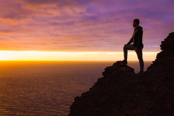 Visionary man standing on top of cliff edge staring at colorful sunset by the sea in Gran Canaria Silhouette of person witnessing unique twilight from mountain top. Successful, entrepreneur concepts conquering adversity stock pictures, royalty-free photos & images