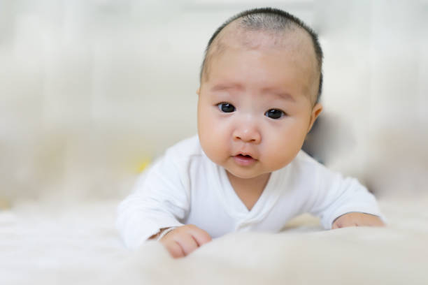 Asian baby learning to crawl and playing with colorful toy in white sunny bedroom. An Adorable Cute laughing child crawling on a play mat. Nursery interior, clothing, and toys for little kids stock photo