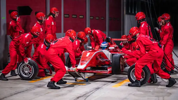 A large group of people in red uniforms at a servicing an open-wheel race car during a pit stop.