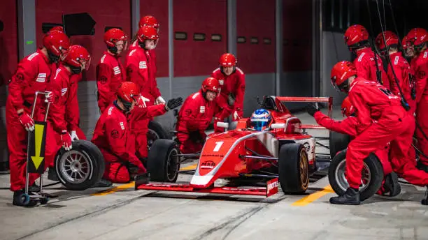 Pit crew in red uniforms preparing to service a red formula race car and change it's tires.