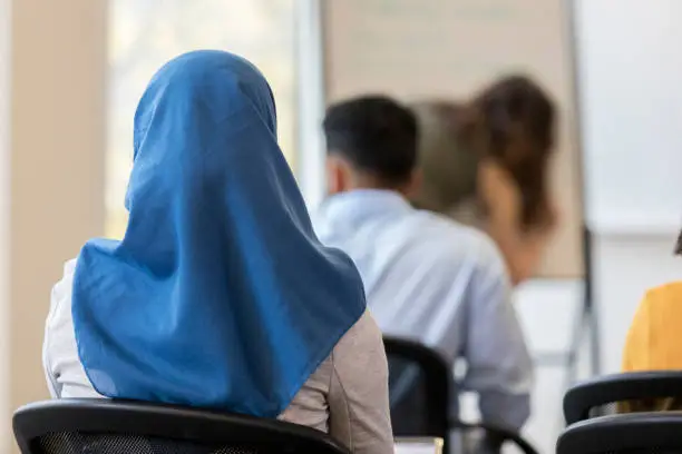 In this closeup rear view, an unrecognizable woman wearing a hijab sits in a chair in a classroom.  Other classmates sit in front of her.