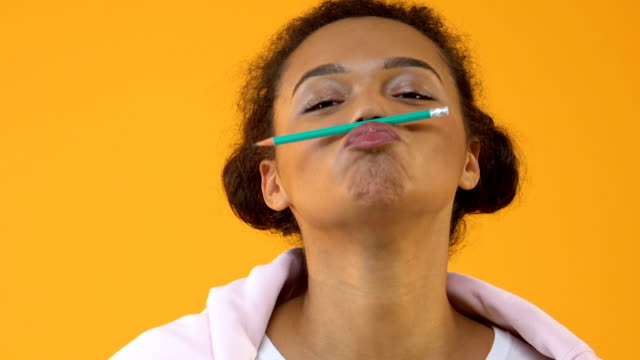 Positive african teenager holding pencil between nose and lips, mustache joke
