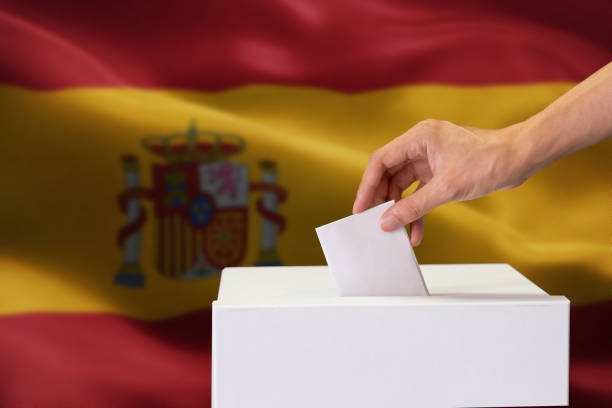 istock Close-up of human hand casting and inserting a vote and choosing and making a decision what he wants in polling box with Spain flag blended in background. 1126041283