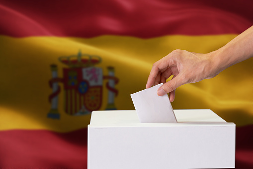 Close-up of human hand casting and inserting a vote and choosing and making a decision what he wants in polling box with Spain flag blended in background