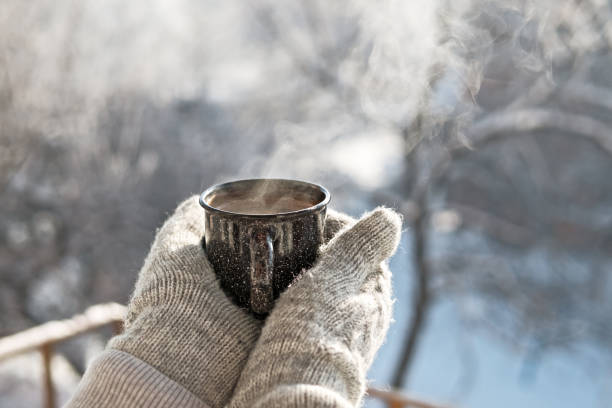 Woman hands in mittens holding a cozy steaming mug or cup with hot cocoa, tea or coffee. Beautiful girl enjoying winter morning or evening outdoors under snowfall. Winter and Christmas time concept