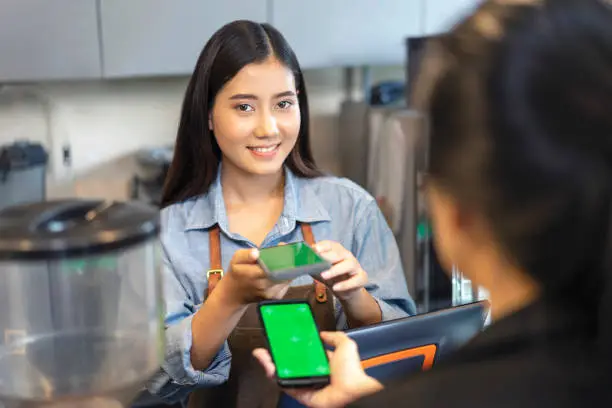 Photo of Customer using her smartphone and nfs high technology to pay a barista for her purchase with phone, transaction and mobile payment, online shop at coffee cafe.
