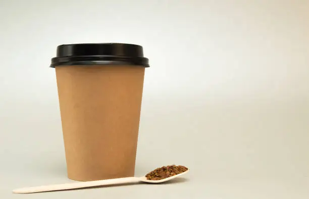 paper cup for coffee with a black lid on a light background, next to it is a wooden spoon with coffee