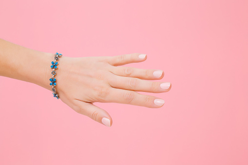 Blue flower bracelet on woman's wrist. Isolated on pastel pink background. Care about hand skin and nails. Closeup.