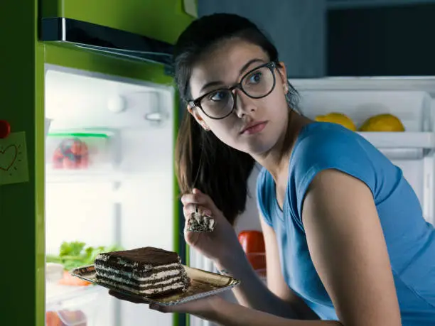 Photo of Woman in the kitchen having a late night snack, she is taking a delicious dessert from the fridge, diet fail concept