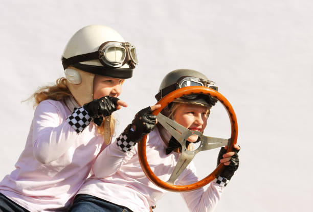 Twin sisters play race car driver sitting in their imaginative sports car.They play happily wearing vintage helmets, goggles and use anold-timer steering wheel they found in the garage.