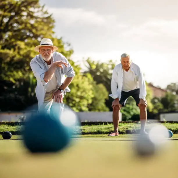 Elderly man playing boules in a playground with his playmate standing in the background. Old man in hat throwing a boules in a lawn with blurred boules in the foreground.